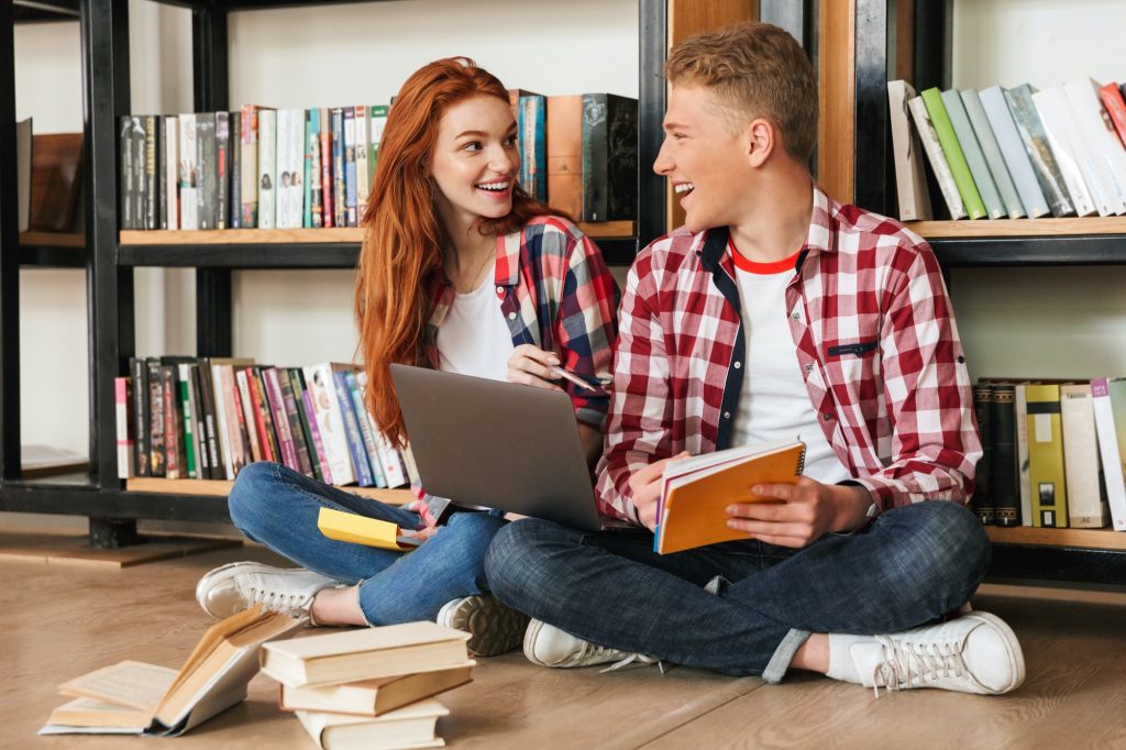 Cheerful teenage couple sitting on a floor at the bookshelf