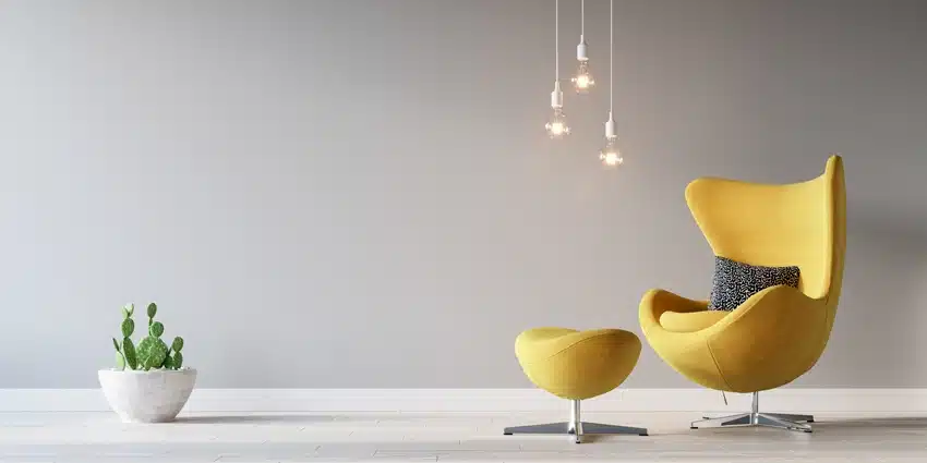 Sculpted yellow chair and ottoman with hanging lightbulbs and a potted cactus against a gray wall, showing many of the top 5 home décor trends in 2021