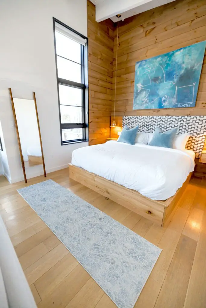 Wood platform bed with white duvet, white pillows, and blue throw pillows against an upholstered headboard and wood paneled walls.