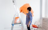 Pretty smiling middle-aged woman painting interior wall of home with paint roller. Redecoration