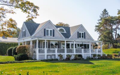 Two-story light gray Cape Cod home with wrap-around front porch in white trim.